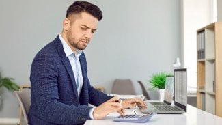 Muž v obleku sedící za kancelářským intenzivně počítá na kalkulačce
