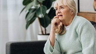 Seniorka sedí na gauči, tváří se zamyšleně, jedním prstem si sahá na ret